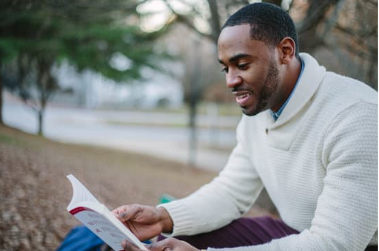 A Black man smiles as he reads a book in a park