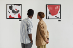 Two people dressed in semiformal apparel are seen looking at two pieces of art comprised of black and red blob-form abstract pieces.