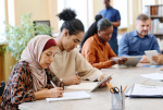 Four students of different ethnicities sit at a table studying. The student closest in the foreground is a Muslim Hijabi woman.