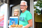 Chef John Beilfuss stands next to his wife, Morgan Beilfuss. He is white with black frame glasses, white short hair, and is wearing a green tee shirt that says "Hola" in capital blue text. He holds a plate of their award-winning fried chicken. His wife Morgan stands to his right side, She is white with long, straight blonde hair and bangs. She is holding their MLive award for the best fried chicken in Michigan.