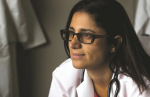 A picture of Dr. Mona looking into the distance. She has dark, straight brown hair that is shoulder length, and thick brown acrylic glasses. She wears a white coat and a stethoscope around her neck.