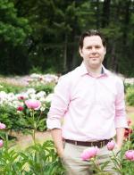 A picture of Kiel Phegley, a tall white man with short brown hair. He is wearing a pink button down shirt with the sleeves rolled up to his elbows, a brown belt, and khaki pants. He stands in a field with bright pink flowers and a forest behind him.