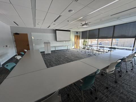 Photo of large room with tables and chairs in a U-shape, podium, and ceiling mounted projector screen.