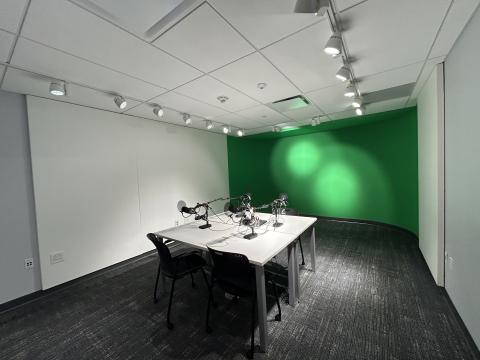 Photo of digital studio with table and four chairs, podcasting board, four microphones and green screen.